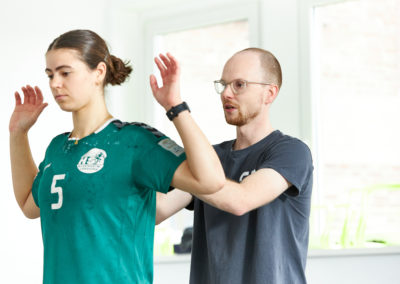 Physiotherapeut Malte Klett behandelt Sportlerin am oberen Rücken welche mit Händen nach oben vor ihm steht - Sport & Schmerz Campus