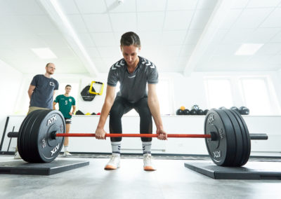 Personal Trainer Malte Klett leitet Sportlerin an der Langhantel an in den Trainingsräumen des Sport & Schmerz Campus, während andere Sportlerin zuschaut
